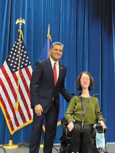 Naomi poses for a photo with a man in a suit. Both are in front of a blue velvet curtain and an American flag.