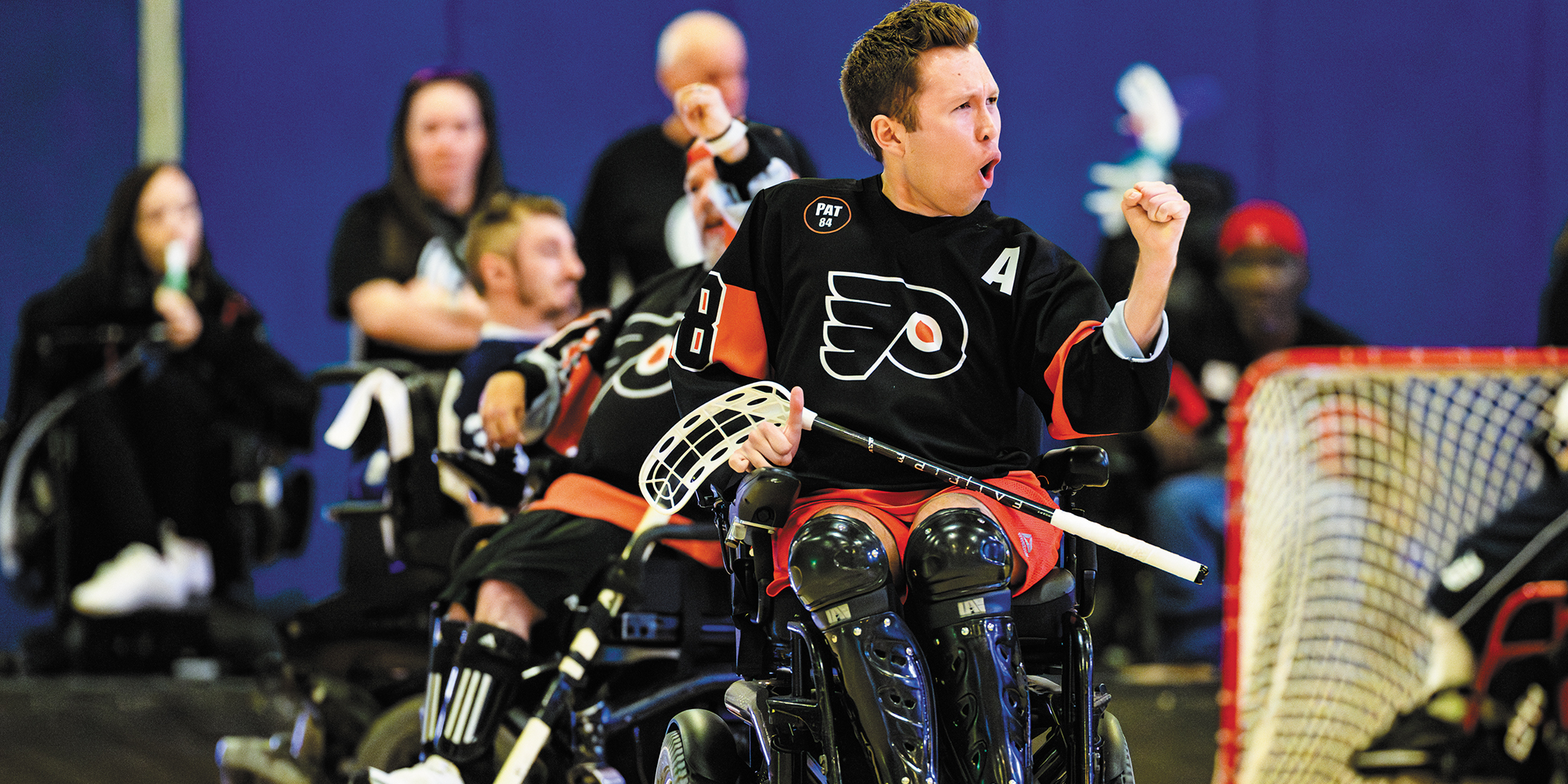 Action shot of Jake Saxton pumping his left fist with a triumphant look on his face and his mouth open in a yell. He is in a power wheelchair wearing a hockey jersey and knee and shin guards, with a power hockey stick resting in his right hand.