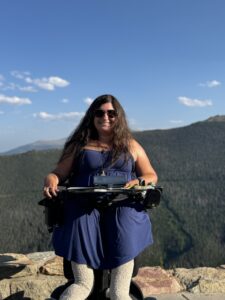 Lyza Weisman, a law student who lives with spinal muscular atrophy, sits in her power wheelchair in front of a scenic overlook with a view of forested mountains.