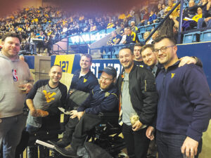 In front of stadium bleachers, Eric Arnold sits in an elevated power wheelchair in the midst of a group of men, all wearing WVU shirts or hats.