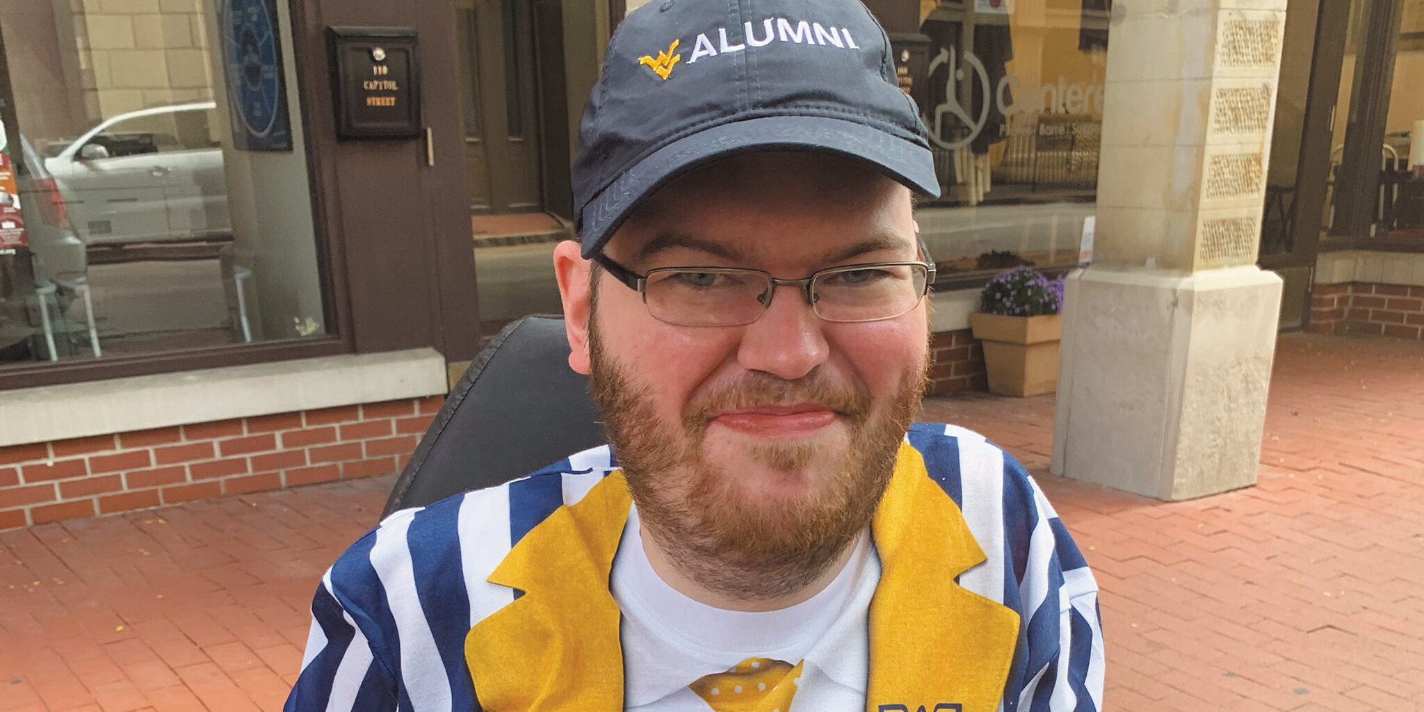 Closeup of Eric Arnold, a light-skinned man with a closely trimmed brown beard and wire-rimmed glasses, wearing a baseball cap that says WVU Alumni.