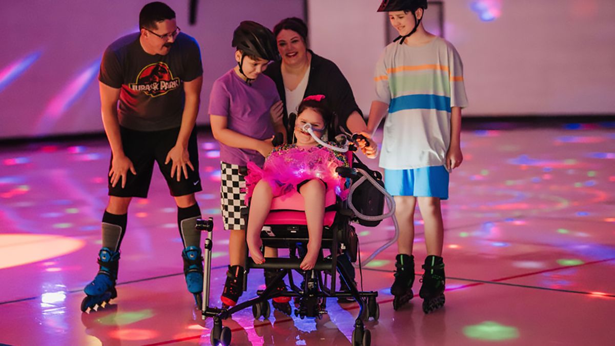 Aubrey’s family gathers around her, smiling, on the wooden floor in a skating rink. The colored lights cast a pink glow over Aubrey in her wheelchair and her family members standing in roller skates.