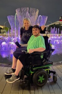 Christ West stands next to her son Zach, who sits in a power wheelchair, in front of a fountain lit with purple and green lights at night.