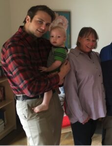 Gavin Beeker stands in the foreground, holding a toddler to his chest, with is mom Ranae to his side.