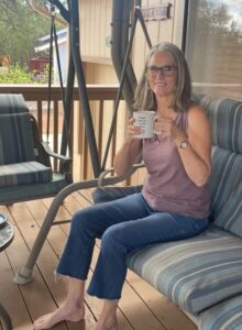 Karen Clough sits on a patio swing holding a cup of coffee, dressed for comfort in a tank top and jeans, with bare feet.