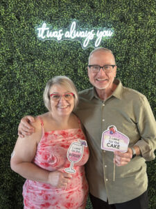 Lillie and Ken Cleeton pose in front of a wall covered in green shrubbery with a neon sign that says “it was always you.” Lillie holds a small sign shaped like a wine glass that says “I came for the booze.” Ken holds a small sign shaped like a slice of cake that says “I came for the cake.”