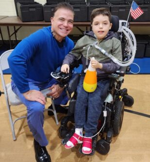 Arturito Estopinan sits in a power wheelchair with one hand on the controller and the other gripping a toy, while his father sits in a chair smiling beside him.