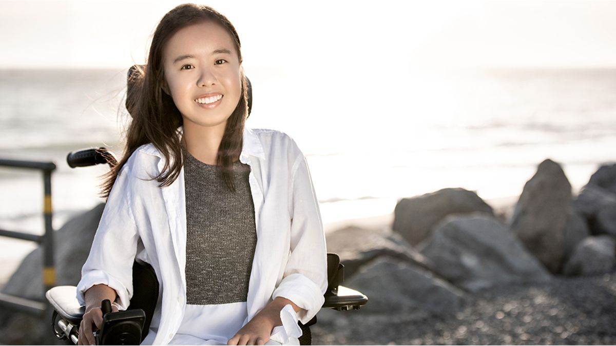 Rachael Bae sits in her wheelchair and smiles at the camera in front of a rocky shoreline bathed in soft light.