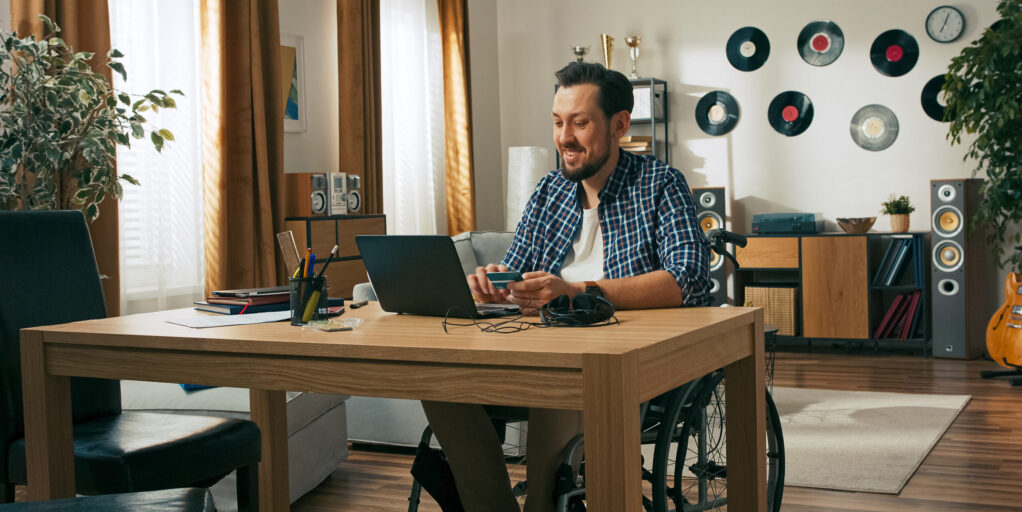 A cheerful man with a disability works remotely in his wheelchair. After completing a task, he closes his laptop and places his card on the table, enjoying the freedom of digital payments.