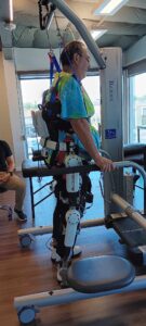 Dwayne Wilson stands in a physical therapy clinic with his torso and legs strapped into an exoskeleton device. He is holding onto the handlebars of a movable frame for balance.