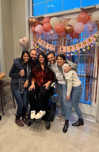 A group of people gather around a young woman in a wheel chair with a Happy Birthday banner in the background