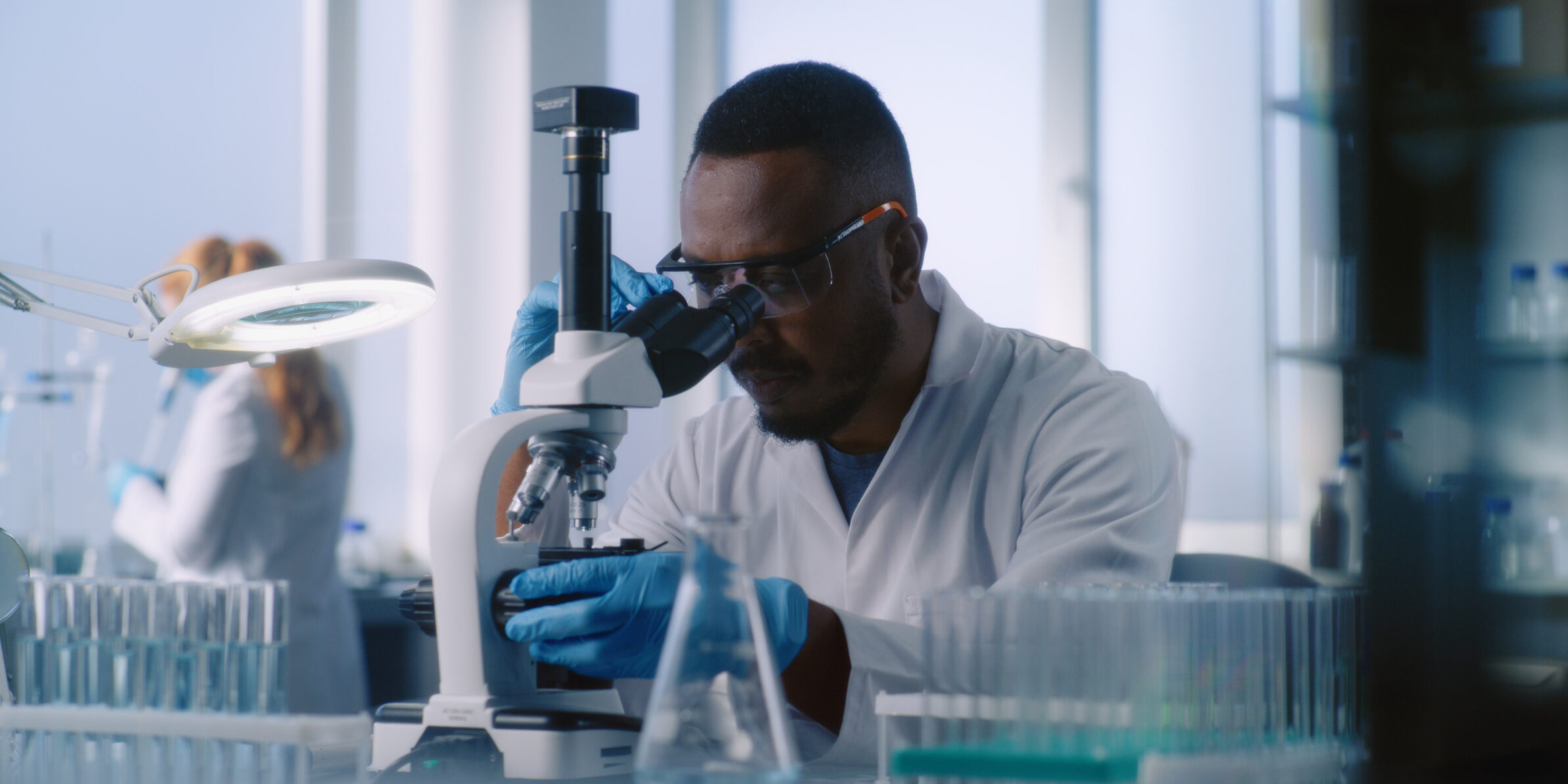 Scientists in white coats work in advanced laboratory. African American microbiologist examines liquid from test tubes with samples under microscope.