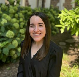 Headshot of Payton Rule, a young woman with brown hair in a black shirt smiling in front of hydrangea bushes