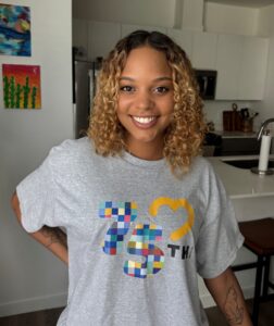 Headshot of Santana Gums, a young woman with curly highlighted hair wearing an MDA t-shirt