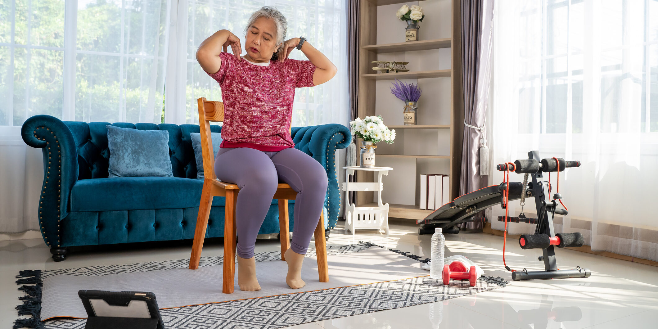 Senior woman sitting on a wooden chair and slowly rotate both shoulders, doing with training exercise online on tablet, workout online from home concept