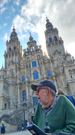 A man in a power wheelchair and baseball cap sits in front of a large cathedral in Santiago, Spain