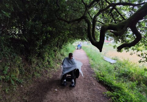 View from behind a man in a power wheelchair traveling on a tree lined dirt path