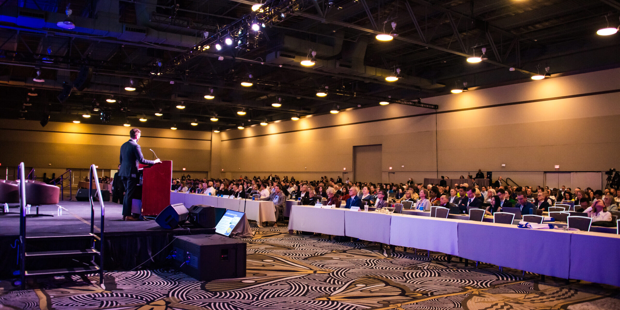 Side view of a conference room full of seated audience members with a man speaking at a microphone