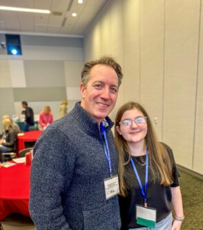 A young woman poses with a man, both wearing name tag lanyards at a conference