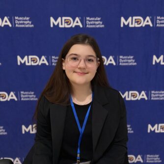 A young woman with dark brown hair and glasses smiles in front of an MDA backdrop