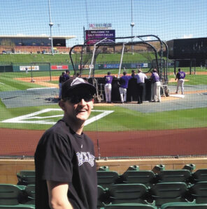 Michael Chamberlain in the fourth row of seats at Salt River Fields stadium in Arizona. Several baseball players are gathered on the field. Michael is wearing a Colorado Rockies baseball cap and T-shirt and is turning away from the field to look at the camera.
