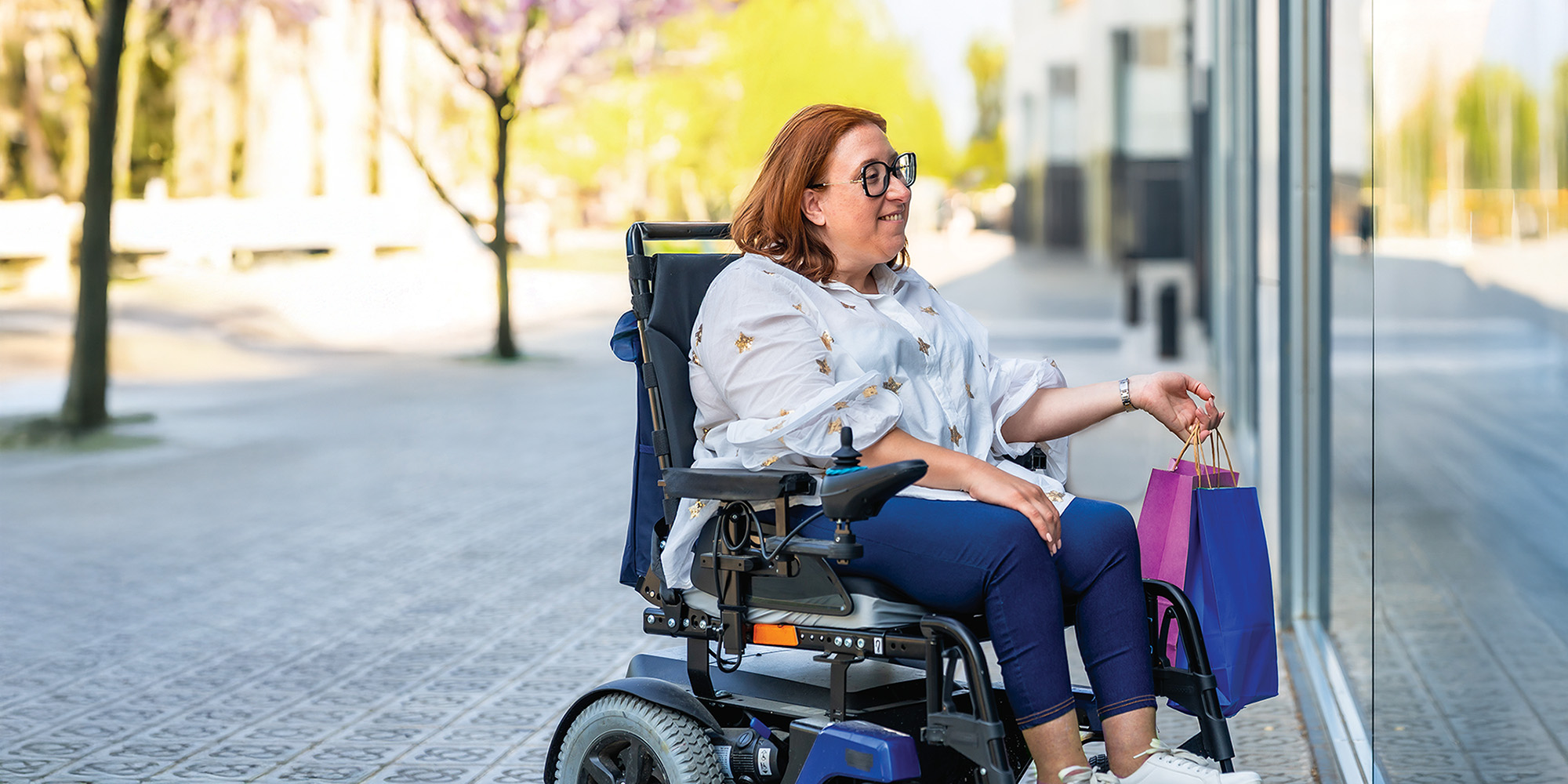 Smiling woman in a wheelchair enjoying the view of a shop window while holding several colorful shopping bags in her hands