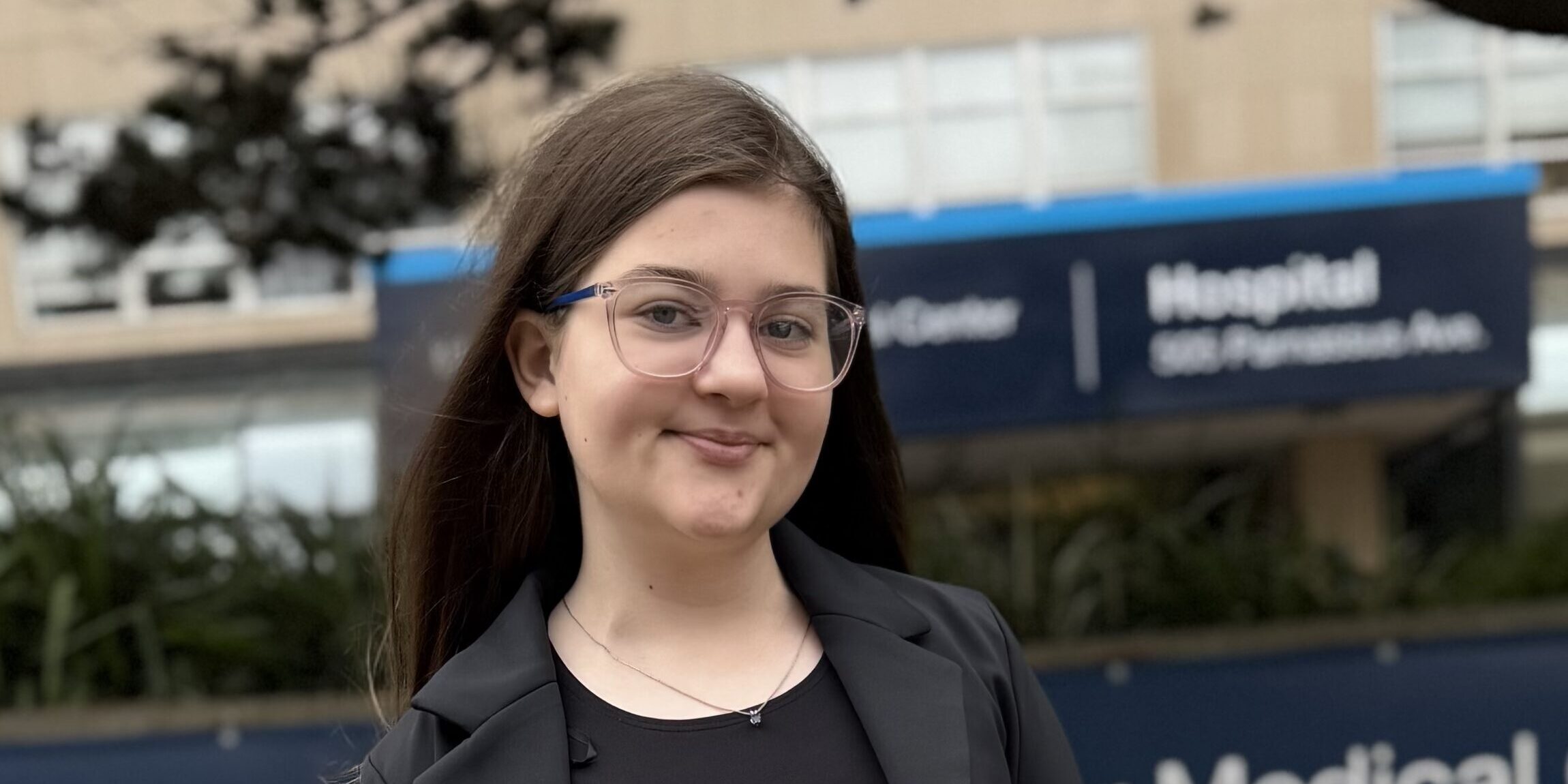 A young woman with dark hair and glasses smiles in front of a hospital