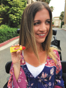 Closeup of Alexa Dectis sitting in a power wheelchair holding up a yellow and red tube of Carmex lip balm.