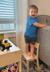 Two-year-old Grayson stands barefoot on a child-sized chair balanced against a couch, with a big, open-mouthed smile at the camera.