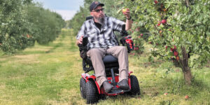 David Daw reaches his left arm up to pick a yellowish apple from a tree in an apple orchard. He is sitting in a red and black power wheelchair with rugged, off-road tires.