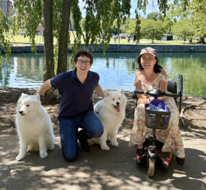 Caroline LeMay, sitting in a mobility scooter, and her husband, holding the collars of two large dogs with thick white coats, pause on a shady path by a lake.