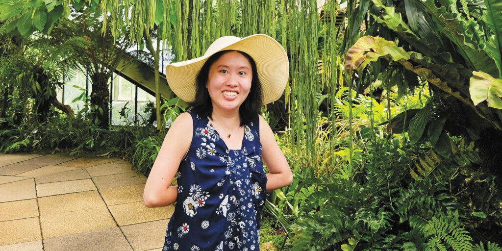 Caroline LeMay smiles at the camera while she stands on a flagstone path in a garden wearing a flower print dress and a straw sun hat.