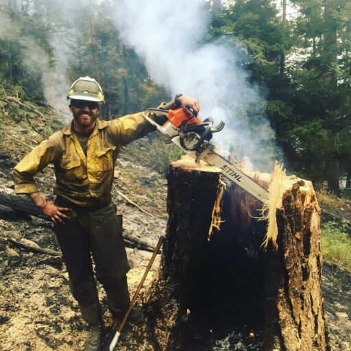 Wildland fire fighter Tyler Long stands in front of a smoking forest fire with a chainsaw and freshly cut tree