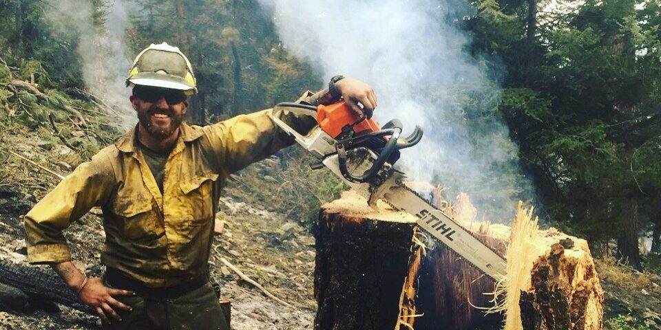 Wildland fire fighter Tyler Long stands in front of a smoking forest fire with a chainsaw and freshly cut tree