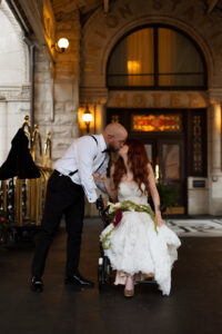 Matt Taylor, dressed in a tuxedo without the jacket, bends down to kiss Fiona Cauley, wearing a white lace wedding dress and sitting in a power wheelchair, with the entrance to the Union Station Hotel in the background.
