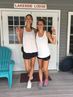 Two young women in white tank tops and black shorts stand in front of a camping lodge