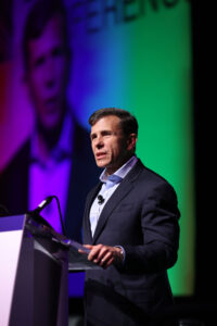 Closeup of John Crowly stands at a podium in the spotlight on a stage.