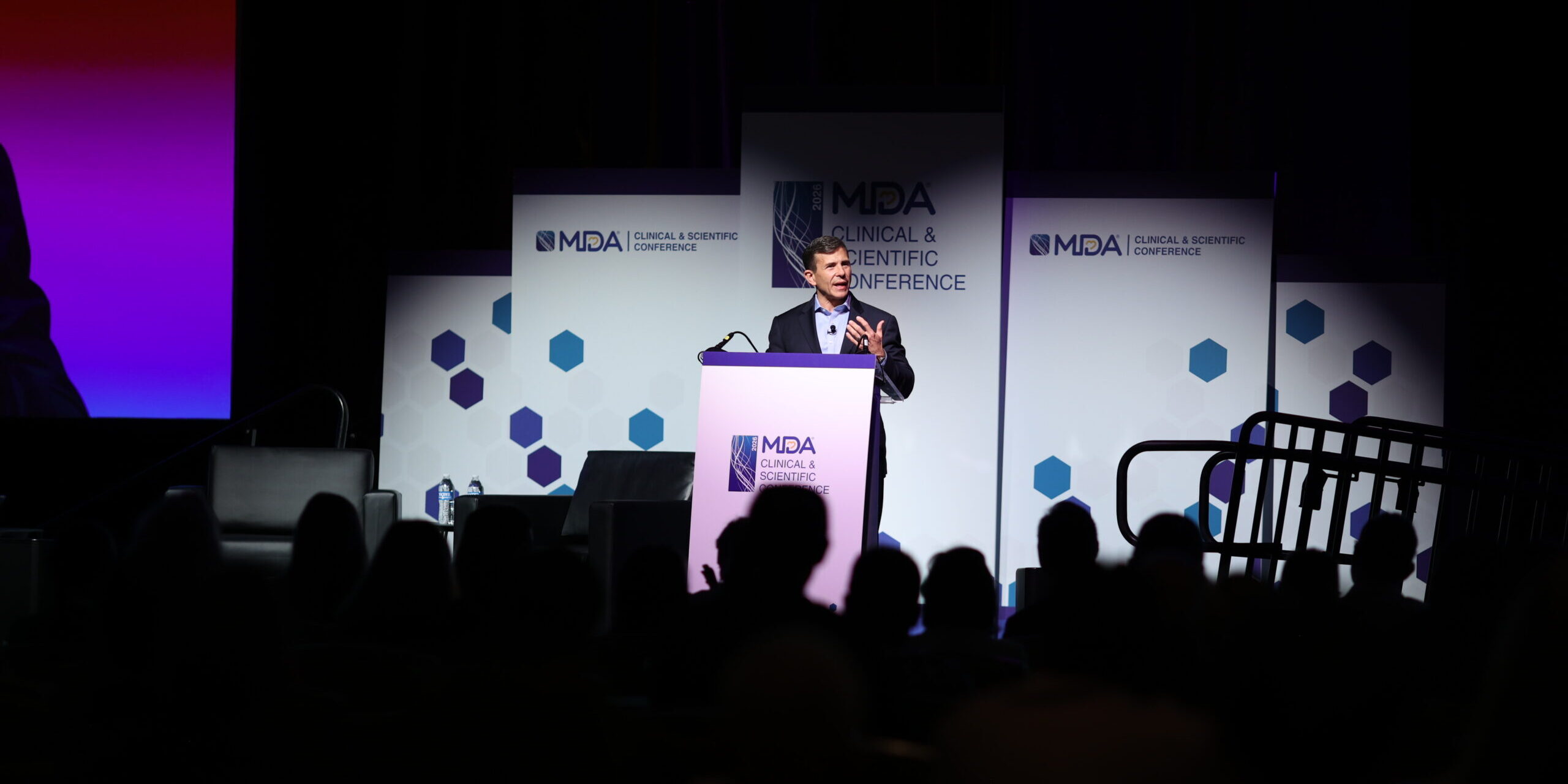 John Crowly stands at a podium in the spotlight in a dark auditorium. Signage on the podium and behind him says MDA Clinical and Scientific Conference.