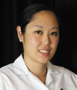 Headshot of Dr. Angela Lek in a white coat smiling against a dark background.
