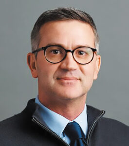 Headshot of Dr. Chet Villa wearing glasses, a tie, and a dark sweater against a gray background