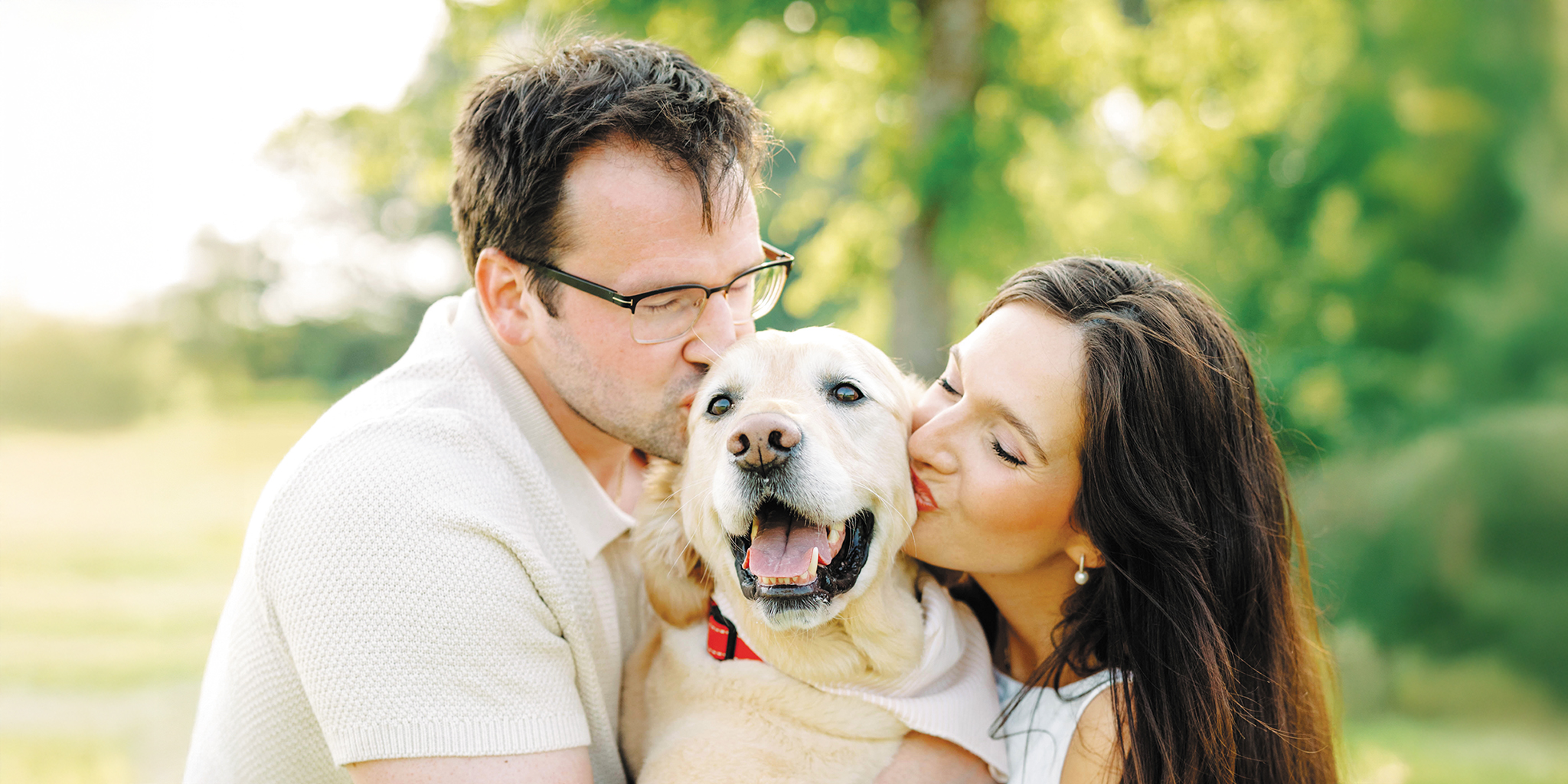 Couple kissing a smiling golden retriever outdoors in a sunny park.