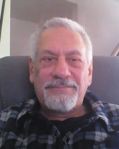 Closeup of Chuck Vrasich sitting in a stuffed chair wearing a flannel shirt.