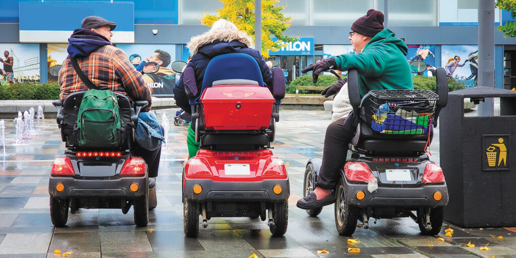 Three people on mobility scooters chatting in a city plaza on a rainy day.