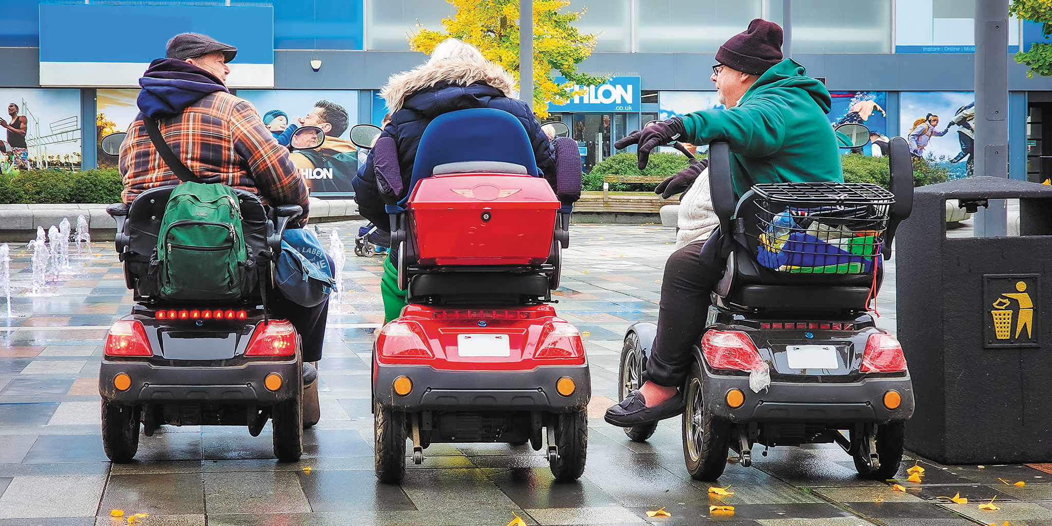 Three people on mobility scooters chatting in a city plaza on a rainy day.