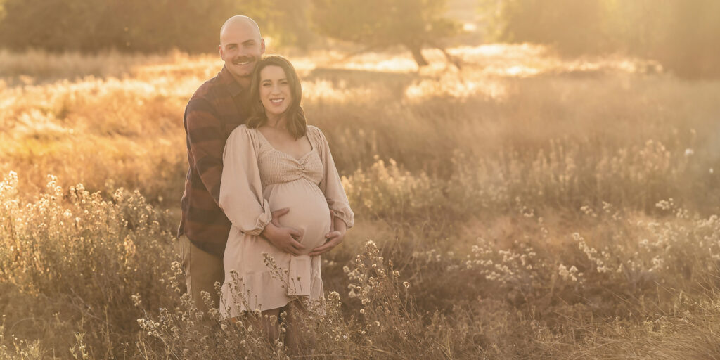 Savannah Jordan and her partner, embracing in a sunlit field, both have their hands lovingly around her pregnant belly.