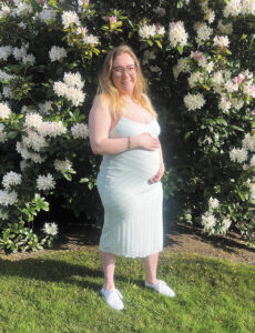 Jessery Picard, while pregnant, stands in a white dress in front of blooming white flowers outdoors.