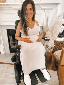 Megan DeJarnett, while pregnant, sits in her power wheelchair in front of a white fireplace, with her hands lovingly on her belly.