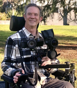 Paul Miller in a wheelchair outdoors with adaptive birding equipment attached to his wheelchair on a swivel arm.