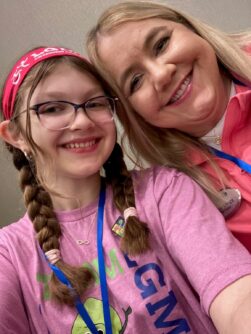A young girl with glasses and two braids smiles next to her mother