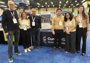 A group of six people stand beside the Cure ADSSL1 conference booth in an exhibit hall.